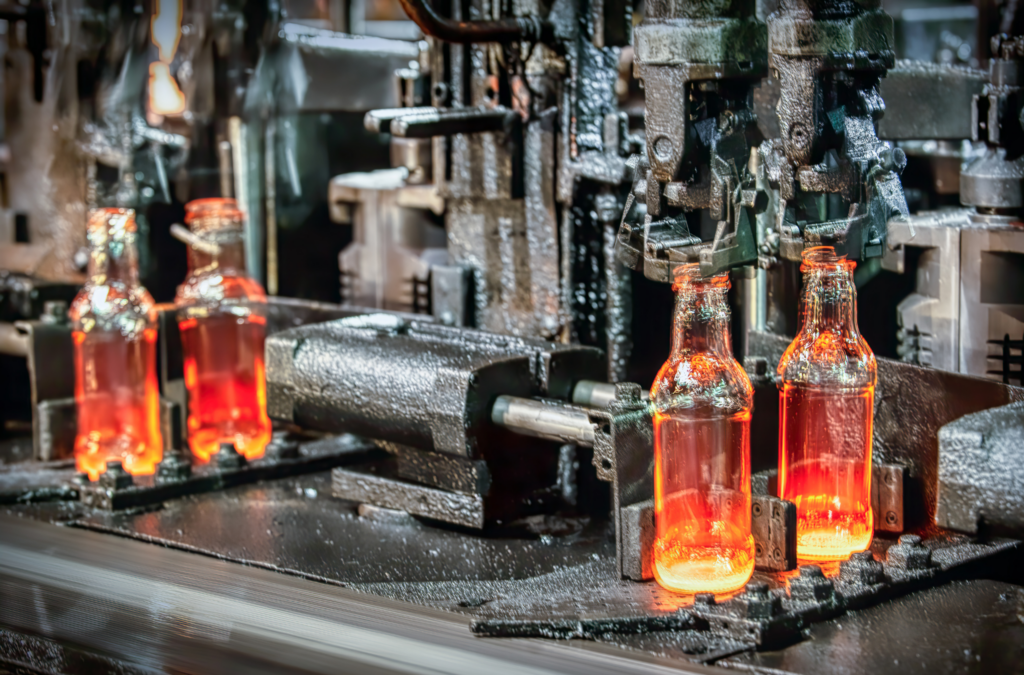 Glowing orange glass bottles being formed by automated machinery on a high-temperature industrial glass manufacturing production line.
